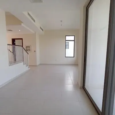 This image captures a spacious and inviting room in a Mira Oasis villa, available for rent. The room is characterized by its clean white walls and tiled floors, complemented by a large window that bathes the space in natural light. A striking feature is the staircase with a sleek white railing leading to the partially visible second floor. Redlac Real Estate Dubai offers this charming 3BR property, combining modern aesthetics with cozy living—ideal for those seeking a serene and bright home environment. Discover more about our exclusive listings and find your perfect oasis with Redlac Real Estate.