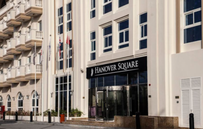 The image showcases a striking multi-story building adorned with a Hanover Square sign, exemplifying modern architecture with its sleek balconies and expansive windows. This elegant structure houses chic studio apartments, perfect for those seeking stylish urban living. Flags gracefully flutter above, while a car parked nearby completes the dynamic city scene. Experience the allure of sophisticated living by exploring Hanover Square and discover your dream home with Redlac Real Estate in Dubai – where luxury meets lifestyle.