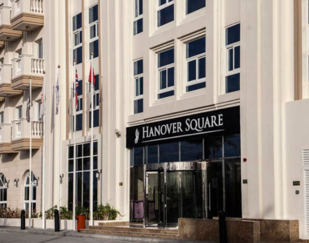 The image showcases a striking multi-story building adorned with a Hanover Square sign, exemplifying modern architecture with its sleek balconies and expansive windows. This elegant structure houses chic studio apartments, perfect for those seeking stylish urban living. Flags gracefully flutter above, while a car parked nearby completes the dynamic city scene. Experience the allure of sophisticated living by exploring Hanover Square and discover your dream home with Redlac Real Estate in Dubai – where luxury meets lifestyle.