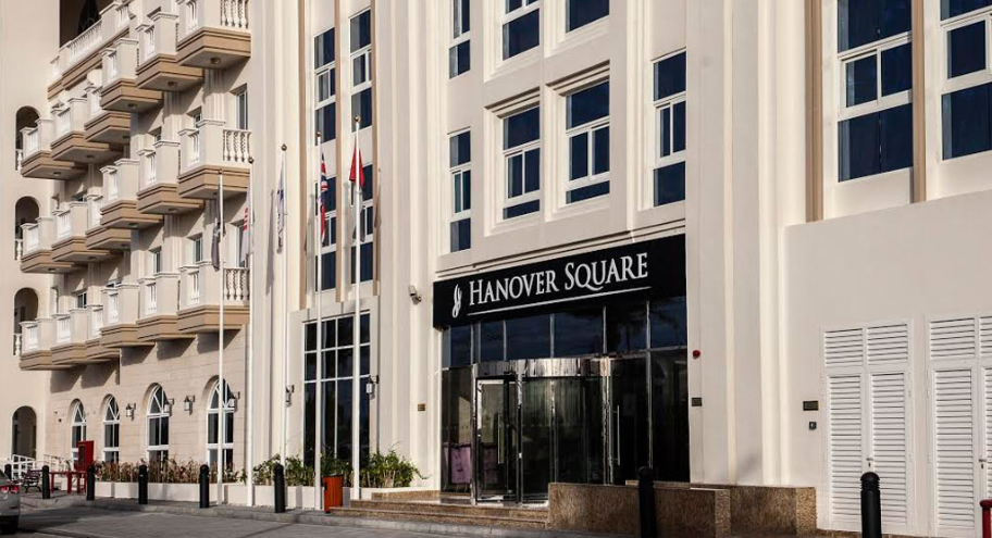 The image showcases a striking multi-story building adorned with a Hanover Square sign, exemplifying modern architecture with its sleek balconies and expansive windows. This elegant structure houses chic studio apartments, perfect for those seeking stylish urban living. Flags gracefully flutter above, while a car parked nearby completes the dynamic city scene. Experience the allure of sophisticated living by exploring Hanover Square and discover your dream home with Redlac Real Estate in Dubai – where luxury meets lifestyle.