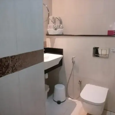 This contemporary bathroom, part of a beautifully furnished studio apartment, showcases a sleek design with features including a wall-mounted toilet and convenient bidet spray. A small white trash bin complements the setup, while the sink is paired with a mirrored cabinet above. Towels are neatly displayed on an organized shelf. Decorated with beige tiled walls enhanced by a stylish border, this space epitomizes modern comfort. Ideal for those seeking rental options in JVC, explore such premium listings through Redlac Real Estate in Dubai for unparalleled living experiences.