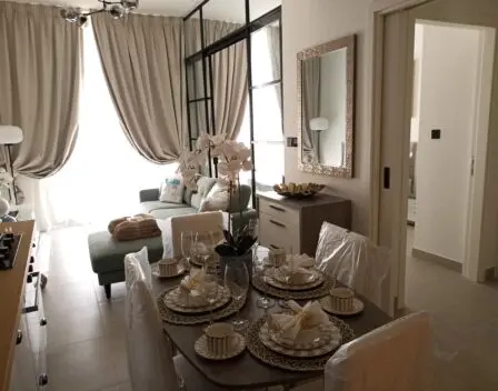 The image showcases a stylish modern apartment interior, highlighting a dining table elegantly set for four. Adjacent to this is a cozy living area with a chic green sofa complemented by large, soft curtains. A wall-mounted mirror beautifully reflects the warm ambient lighting, enhancing the inviting atmosphere. A doorway hints at additional spaces within the home. Discover such exquisite living spaces in Dubai with Redlac Real Estate, where luxury meets comfort in every detail.