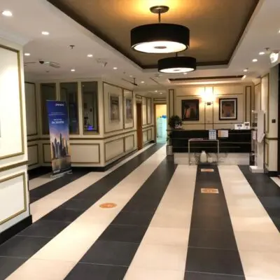 Step into contemporary elegance at a modern hotel lobby in Arjan Dubailand, where stylish decor meets sophistication. The walls showcase framed artwork that complements the chic environment, while the combination of dark and light tiled flooring adds depth to the space. A sleek reception desk provides a welcoming backdrop under circular ceiling lights that gently illuminate every corner. To the left, a bold blue sign catches your attention. Discover such exquisite properties with Redlac Real Estate, your gateway to luxurious living in Dubai. Whether youre investing or seeking your dream home, Redlac Real Estate offers unmatched expertise and an exceptional portfolio in this vibrant city.