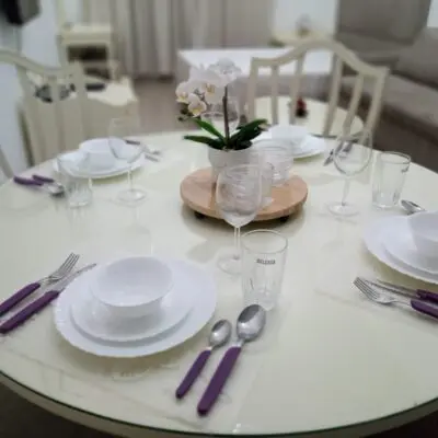 This inviting JVC studio apartment showcases a round dining table elegantly set for four, featuring white plates and bowls alongside clear glasses. The purple-handled cutlery beautifully accentuates the charming potted orchid at the center. In the background, a subtly blurred living area adds to its warm ambiance. Discover such delightful properties and more with Redlac Real Estate, your trusted partner in Dubai for finding your dream home or investment opportunity.