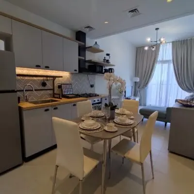 Discover the epitome of modern elegance in this stylish kitchen and dining area, featuring sleek light gray cabinets paired with a warm wooden countertop and a chic backsplash. The inviting dining table is set for four, complete with pristine white chairs, making it an ideal spot for intimate gatherings. In the background, a cozy living room boasts a large window that bathes the space in natural light while elegant lighting fixtures add to the sophisticated ambiance—perfect for hosting one special guest. Experience luxury living at its finest with Redlac Real Estate in Dubai, where attention to detail and premium design meet unparalleled comfort and style.