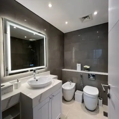 This contemporary bathroom in a stylishly furnished 1-bedroom apartment showcases a sleek design with dark tiled walls contrasting against light floor tiles. It includes a pristine white vanity topped with a round sink, complemented by a backlit mirror, toilet, and bidet. A small shelf adds decorative charm, and bright lighting enhances the welcoming atmosphere. Just steps away, enjoy stunning balcony views. Discover this exquisite living space and more with Redlac Real Estate in Dubai—your gateway to luxurious properties in vibrant locations.