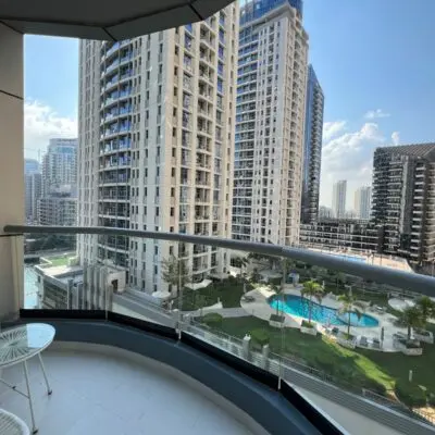 Capture the stunning view from the curved balcony of this elegantly furnished 1BR apartment, where a cityscape of sleek high-rise buildings stretches out before you. Below, a refreshing pool and lush green park offer a serene escape. Relax on the balcony outfitted with two white chairs and a small table under clear blue skies. Discover your dream home with Redlac Real Estate in Dubai, where luxury living meets breathtaking views.