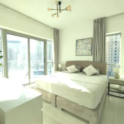 Discover a contemporary 1BR apartment in Dubai, featuring a luminous bedroom equipped with a spacious bed adorned in light gray bedding and complemented by two plush pillows. The room boasts expansive floor-to-ceiling windows that offer stunning city views and lead to a serene balcony. With elegant touches like a chic light fixture, a nightstand complete with a lamp, and tasteful curtains framing the windows, this space epitomizes modern living. Experience luxury and comfort with Redlac Real Estate—your gateway to premium properties in Dubai.