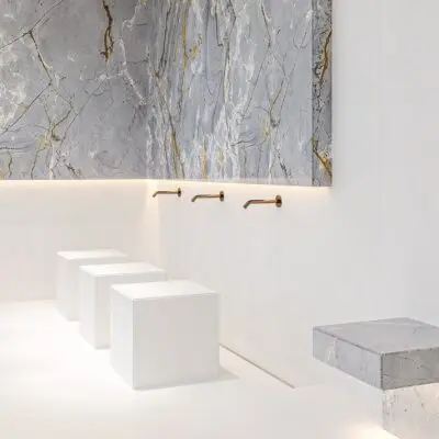 Discover a stylish, minimalist bathroom in this 1-bedroom apartment for sale in Arjan. The space features sleek white cubic stools and a stunning gray marble wall accented with gold veining, creating a luxurious ambiance. Copper faucets elegantly extend from the wall, paired with a floating marble shelf on the right, all beautifully highlighted by ambient lighting. This exquisite property is presented by Redlac Real Estate, your trusted partner in finding premium homes in Dubai.