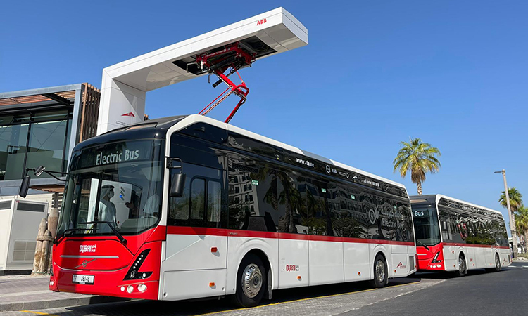 In a vibrant urban setting, two sleek electric buses with distinct white and red designs are parked under a charging station. The scene is bathed in sunlight, accentuating the modernity of the area, which is beautifully juxtaposed with palm trees and contemporary architecture. This image encapsulates the innovative spirit and green initiative ideals synonymous with Dubai. Discover your place in this dynamic cityscape through Redlac Real Estate, where modern living meets sustainability in one of the worlds most iconic destinations.