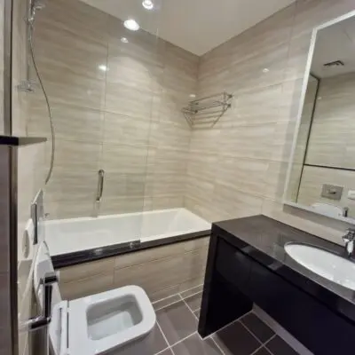 This sleek, contemporary bathroom in Dubai Marina Residences highlights elegant beige tile walls contrasted with a sophisticated dark countertop. It boasts a luxurious bathtub with a glass screen, a wall-mounted showerhead, a toilet, and a spacious mirror above the sink. The floors grid pattern enhances the refined ambiance of Dubais luxury lifestyle. Discover more exceptional properties like this with Redlac Real Estate, your trusted partner in Dubai for finding the ultimate in luxury living.