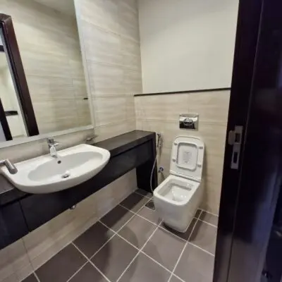 This contemporary bathroom captures the sleek elegance often found in Dubais ultra-modern homes, with its beige tile walls and dark gray floor tiles. It showcases a chic white wall-mounted sink, a large mirror, and a streamlined white toilet with a concealed tank. The dark brown door frame adds an extra touch of sophistication to the space. Discover more stunning properties like this by exploring Redlac Real Estates premier offerings in Dubai.