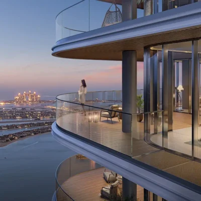 A person gazes at a coastal cityscape from Bayviews curved glass balcony as dusk settles. The vibrant sunset paints the sky, while distant islands and sparkling skyscrapers captivate the eye. Below, warm lights illuminate the bustling streets. Discover such breathtaking views with Redlac Real Estate in Dubai, where luxury living meets stunning vistas and unparalleled lifestyle experiences.