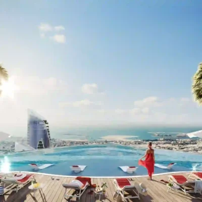 A woman in a striking red dress gazes over a stunning cityscape and ocean from an infinity pools edge under a clear blue sky. Luxurious lounge chairs and palm trees enhance the serene atmosphere, offering an idyllic escape. Discover such breathtaking views and unmatched elegance with Redlac Real Estate, your gateway to Dubais finest luxury properties. Embrace exquisite living today.