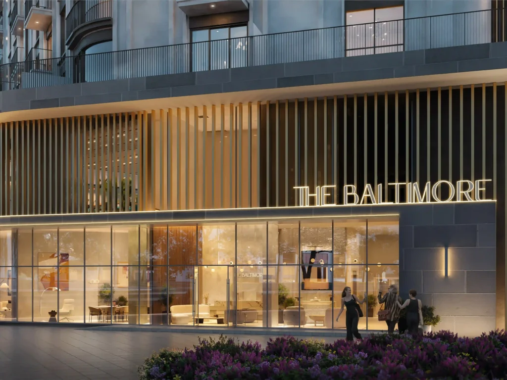 In the heart of Baltimores Town Square, a contemporary building boasts large glass panels and vertical wooden slats. The warmly lit THE BALTIMORE sign radiates luxury as three pedestrians stroll by and purple flowers bloom nearby. Discover similar elegance with Redlac Real Estate in Dubai, where architectural beauty meets opulent living in the worlds most dynamic city.